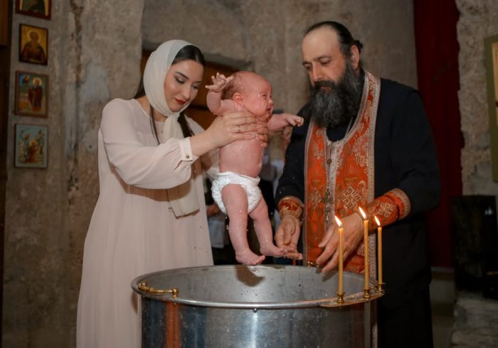 A baby being baptized by a priest and woman in a traditional church setting.