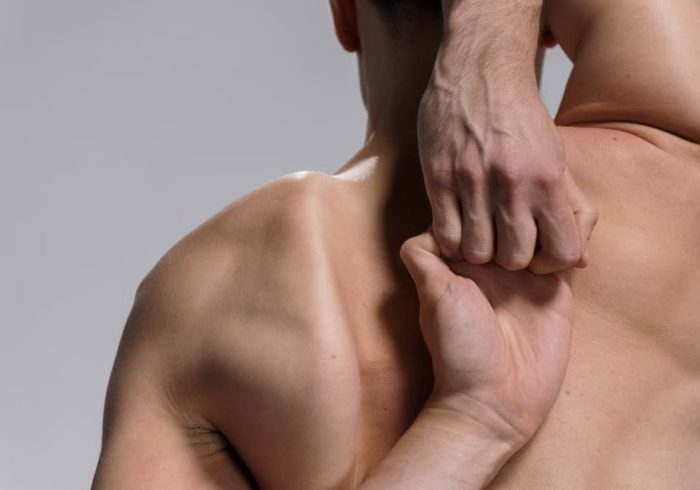 Shirtless muscular man practicing a yoga stretch with hands clasped behind back on a white background.