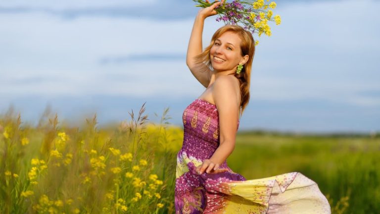 A smiling woman in a vibrant dress holding flowers in a sunny meadow